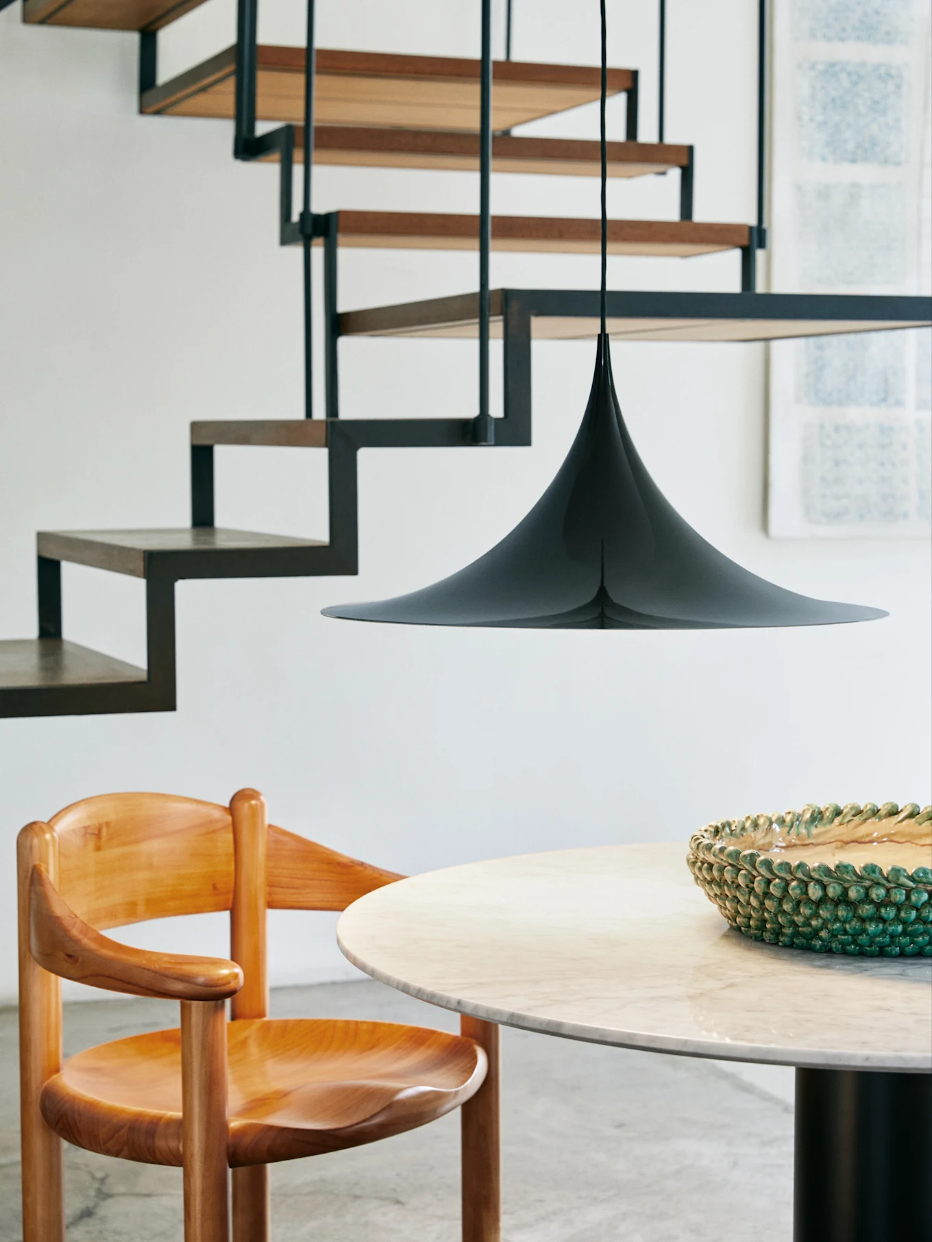 Eine schwarze kegelförmige Pendelleuchte hängt über einem runden Marmortisch und einem Holzstuhl, mit einer modernen Treppe im Hintergrund.