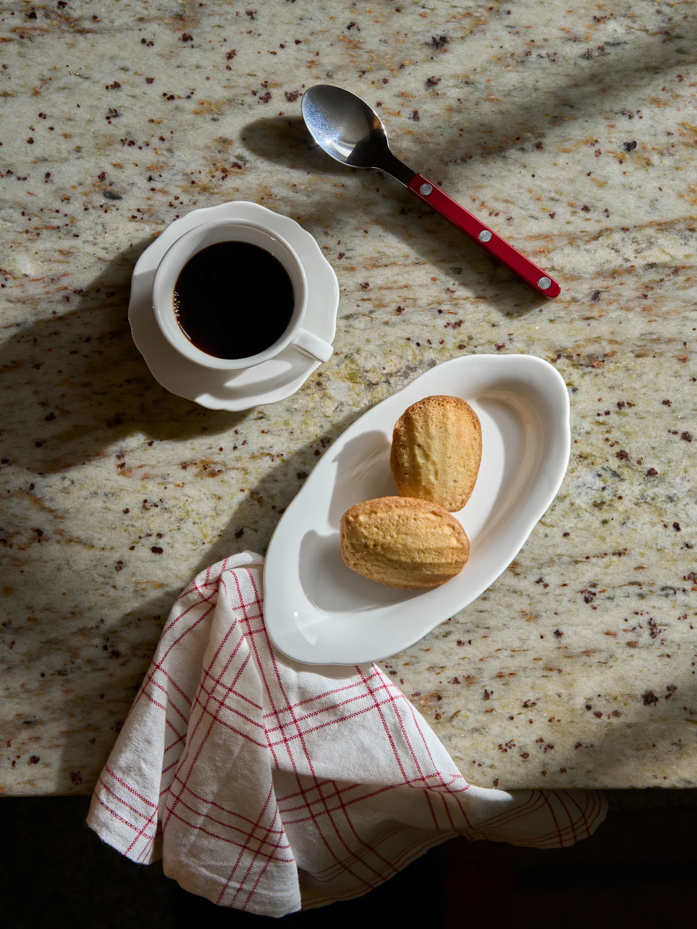 Ansicht von oben: Schwarzer Kaffee in weißer Tasse, zwei Madeleines auf weißem Teller, Löffel und gestreifte Serviette auf Marmorplatte.