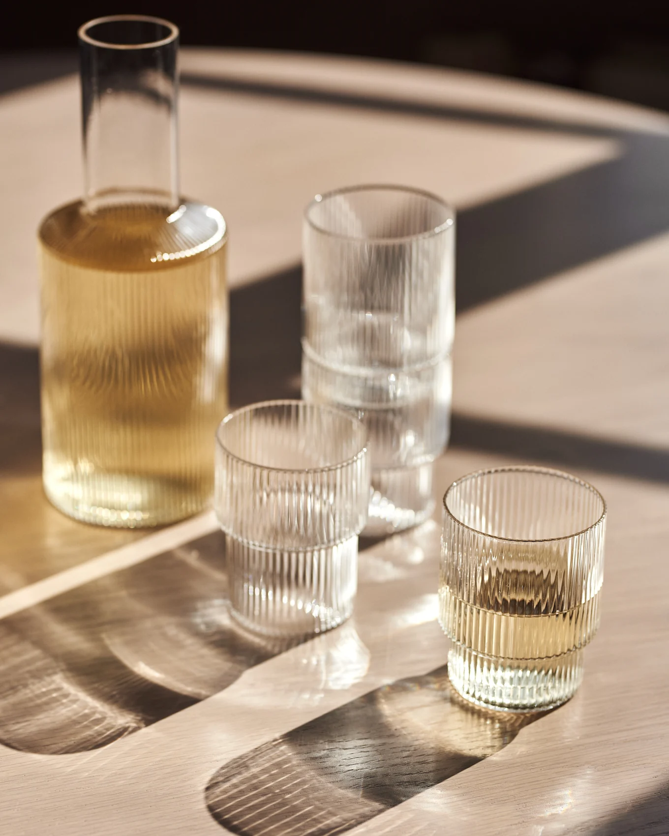Glaskaraffe und gerippte Gläser auf einem Holztisch mit Sonnenlicht.
