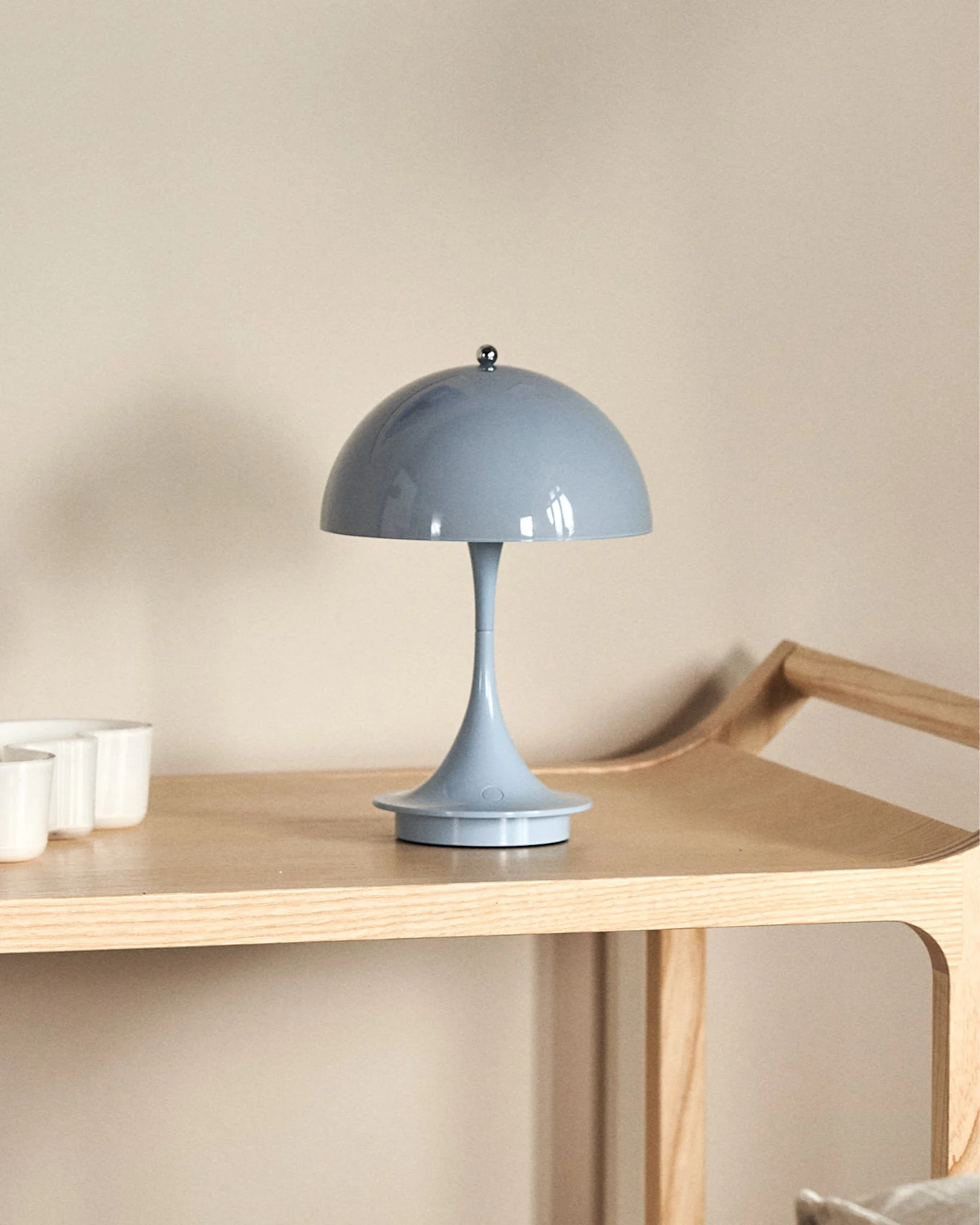 Eine hellblaue, pilzförmige Tischlampe auf einem hellen Holzregal, mit teilweise sichtbaren weißen Tassen links.