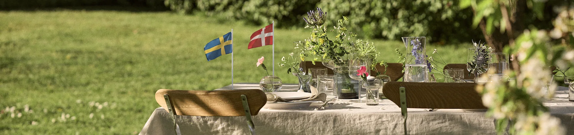 Outdoor-Esstisch mit weißer Leinen Tischdecke, Gläsern, Blumenarrangements und schwedischen und dänischen Flaggen in einem sonnigen Garten.