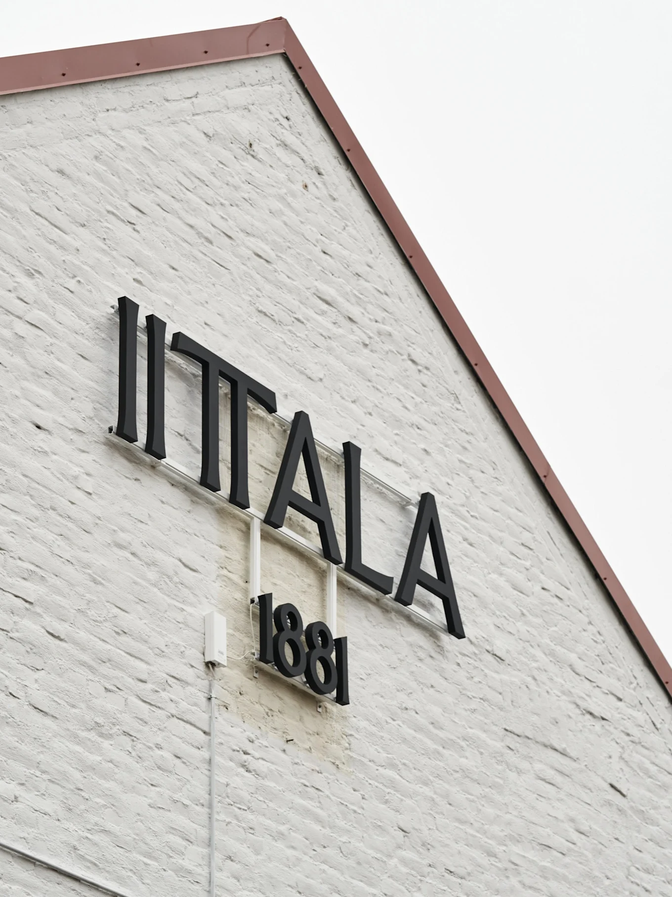 Das Iittala-Logo und '1881' an einer weißen Ziegelwand mit einem rotbraunen Dach.