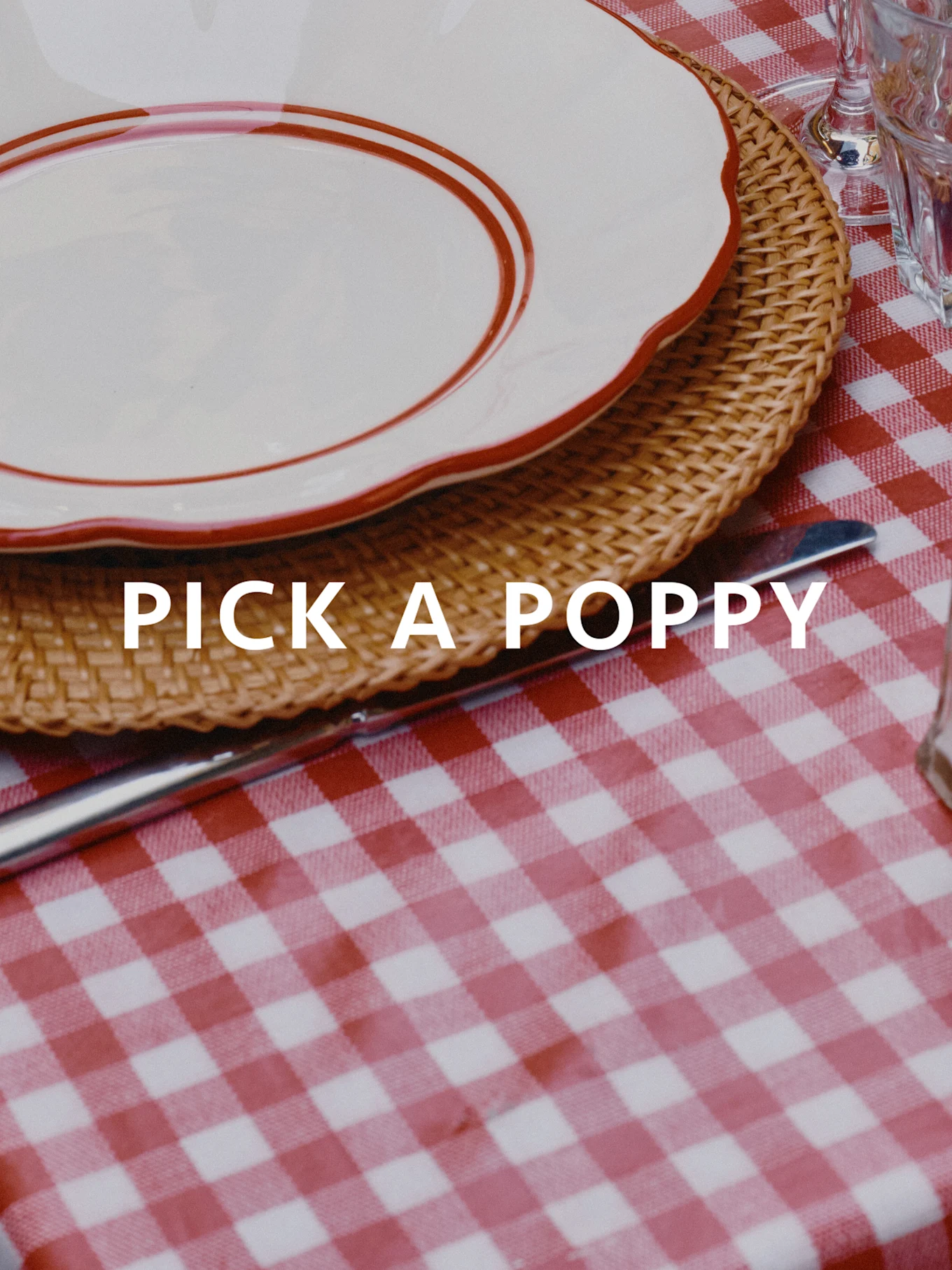 Gedeck mit rot-weiß karierter Tischdecke, geflochtenem Platzset und weißem Teller mit rotem Rand. Text: PICK A POPPY.