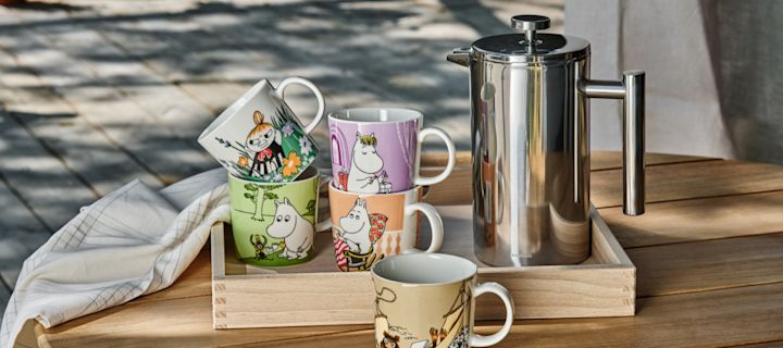 Verschiedene Mumin-Tassen von Moomin Arabia stehen draußen auf einem Tisch, daneben eine Stempelkanne.