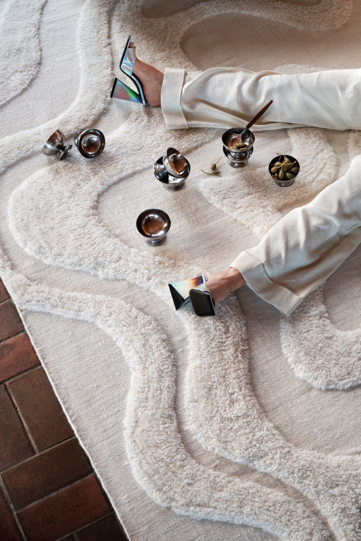 Lernen Sie die beliebte schwedische Marke Tinted Objects kennen: Hier sehen Sie den weißen Wollteppich Norlander auf einem Holzfusboden liegen, darauf mehrere silberfarbene Becher und Frauenbeine in High Heels.