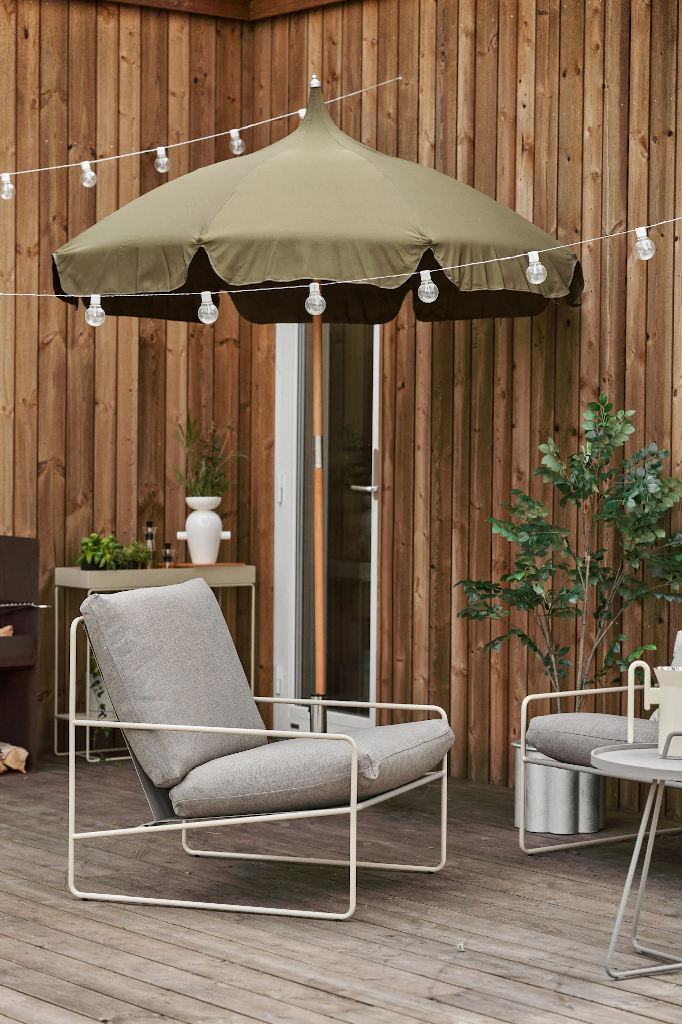 Eine gemütliche Terrasse aus Holz mit Lichterkette, Lull Sonnenschirm und Desert Loungesessel, beides von Ferm Living.