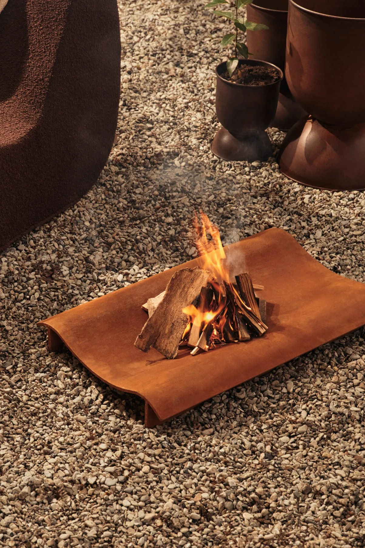 Ein kleines Lagerfeuer brennt auf einem gebogenen, rostbraunen Metalltablett auf einem Kiesuntergrund.