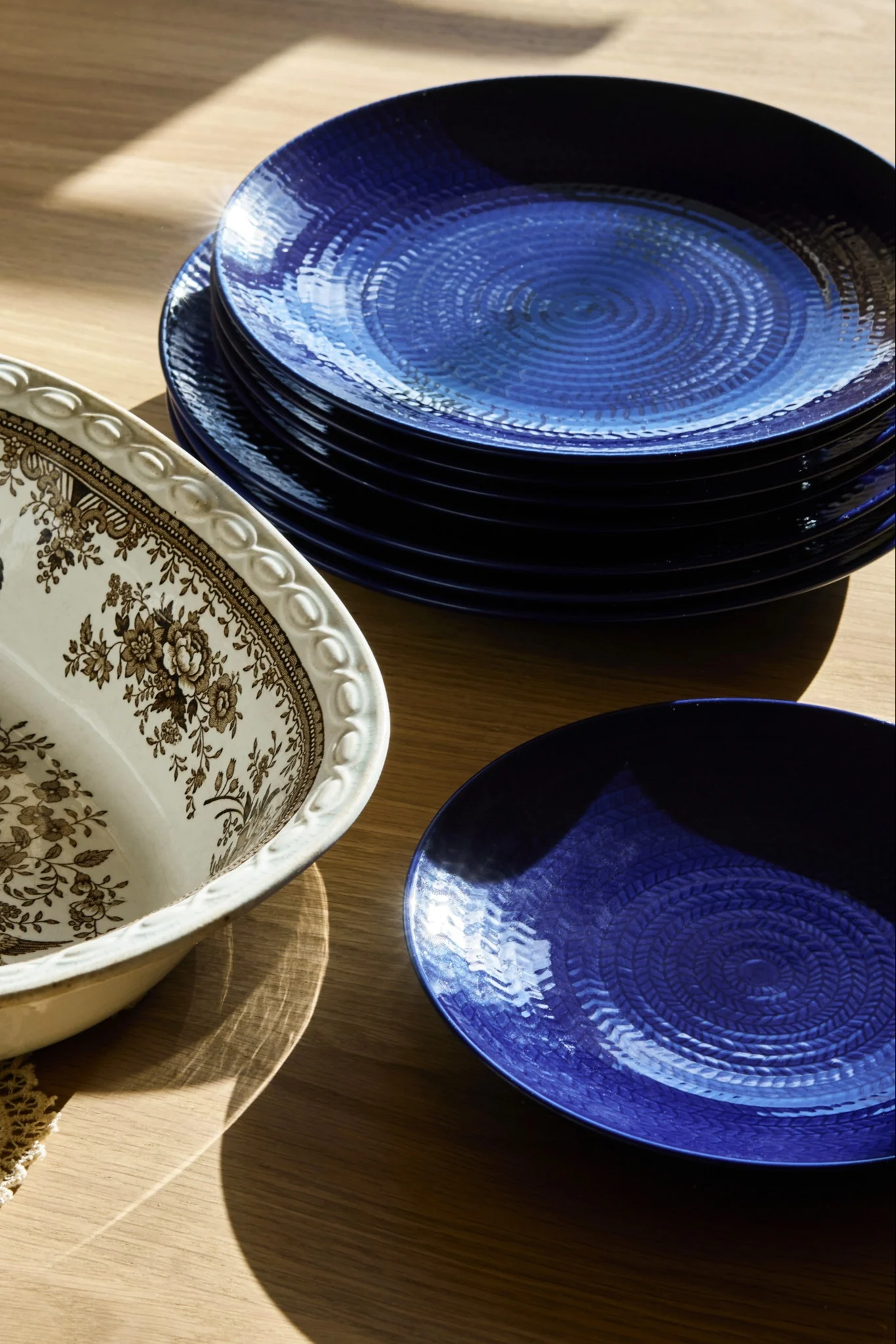 Stapel tiefblauer, texturierter Teller neben einer verzierten weißen Servierschale mit braunem Blumenmuster auf einem Holztisch.