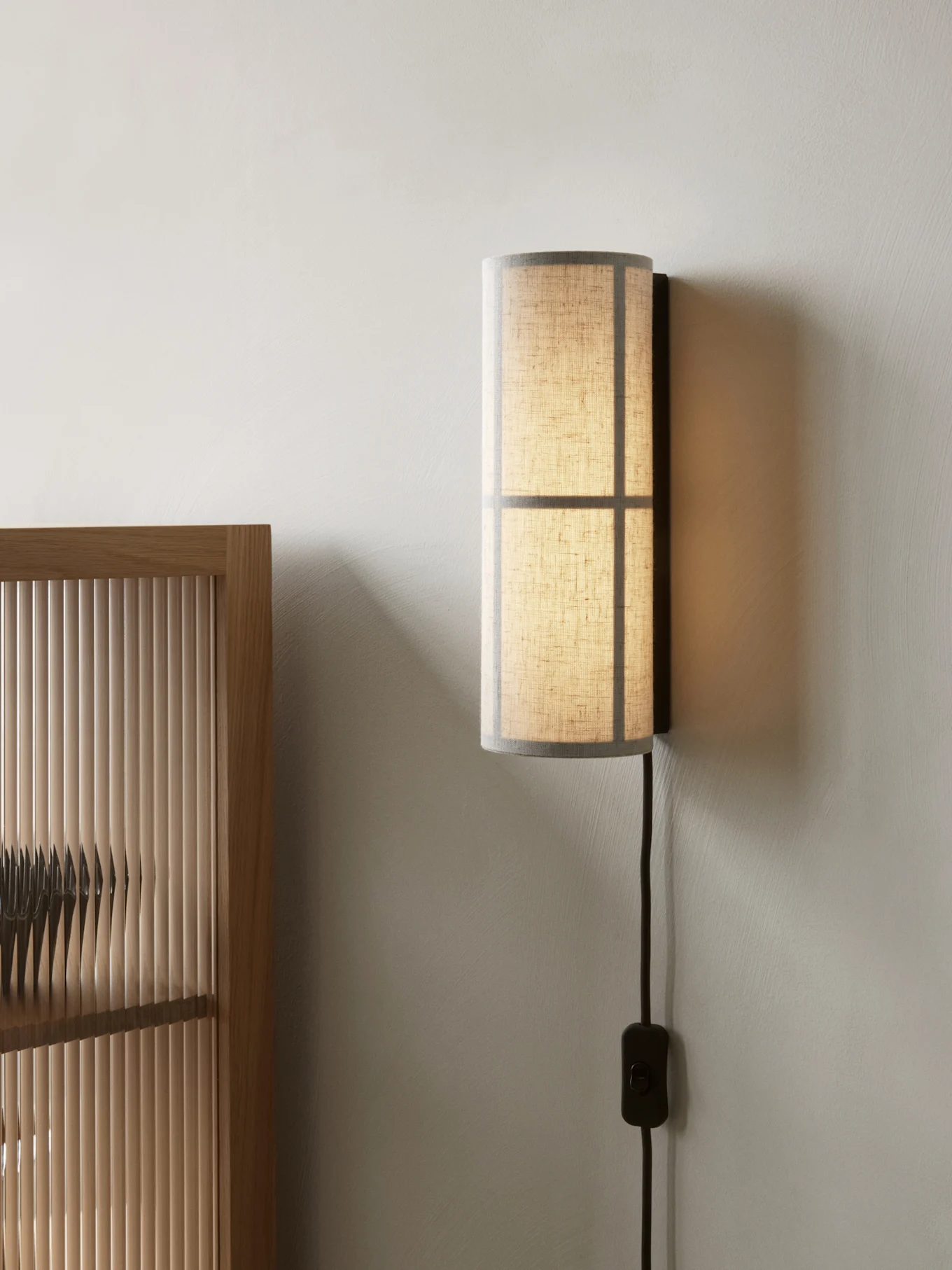 Zylindrische Wandleuchte mit durchscheinendem Stoffschirm, an einer hellen Wand neben einem Holzlamellenschirm montiert.