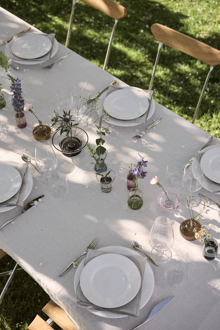 Minimalistische Tischdeko an Mittsommer: Decken Sie den Tisch im Garten mit einer weißen Tischdecke, weißem Geschirr und platzieren Sie mehrere kleine Vasen, wie die aus der Midsummer-Kollektion von Orrefors, wie ein Band in der Mitte.