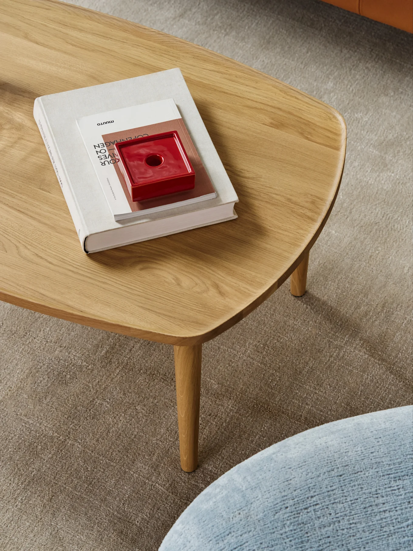 Ein heller Holztisch mit abgerundeten Kanten, darauf ein Stapel Bücher und ein rotes Keramikobjekt, auf einem beigen Teppich.