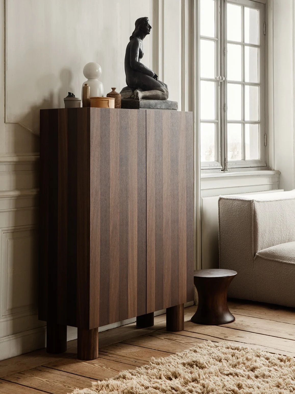 Dunkler Holzschrank mit vertikalen Streifen, gekrönt von einer sitzenden Skulptur, weißer Lampe und Deko, neben einem strukturierten Sofa und dunklem Hocker.