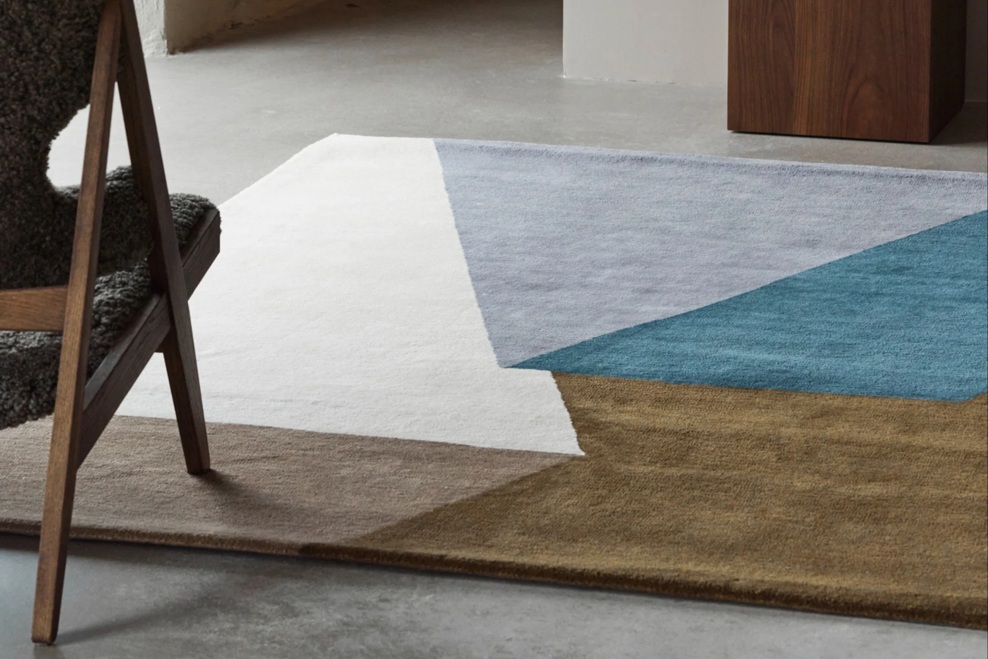 Ein geometrischer Teppich in Weiß, Hellblau, Türkis und Braun auf Betonboden, mit einem Holzstuhl und einer blauen Vase.