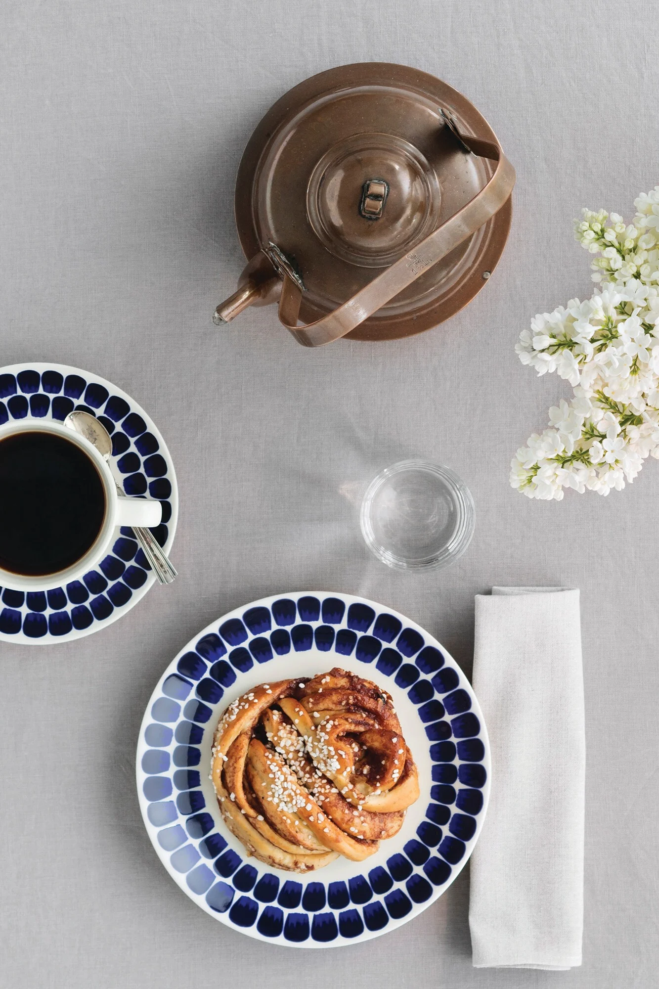 Eine Draufsicht auf einen für eine Fika gedeckten Tisch: Eine Kanne Kaffe, eine Tasse und ein Tuokio Teller von Arabia mit einer Zimtschnecke.