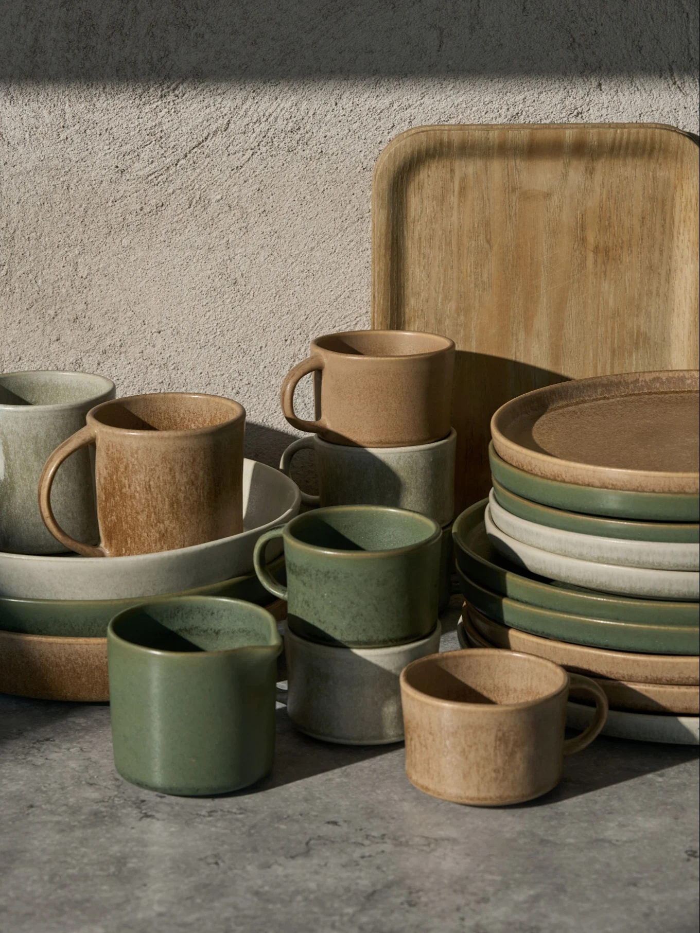 Sammlung grüner, beiger und hellgrauer Keramiktassen, Schalen und Teller mit einem Holztablett auf grauem Untergrund.