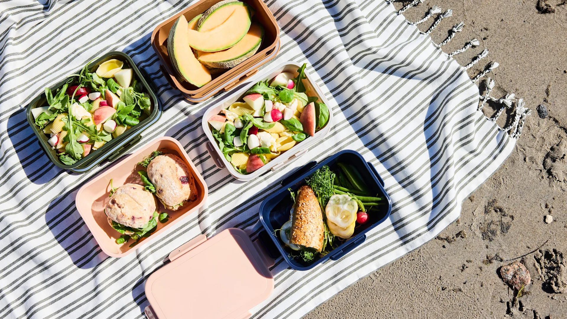 Picknick auf einer gestreiften Decke im Sand, mit mehreren Lunchboxen mit Obst, Nudelsalat und Sandwiches.