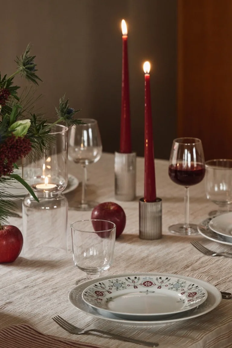 Tischdeko Weihnachten: Ein weihnachtlich gedeckter Tisch mit Geschirr aus der Swedish Grace Winter-Kollektion von Rörstrand, ergänzt durch rote Kerzen und Äpfel als dekorative Details auf dem Tisch.