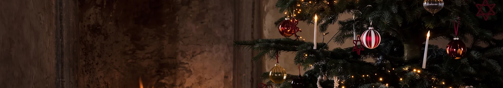 Ein festlicher Weihnachtsbaum mit Ornamenten und einer brennenden Kerze neben einem warmen Kamin mit brennenden Holzscheiten.