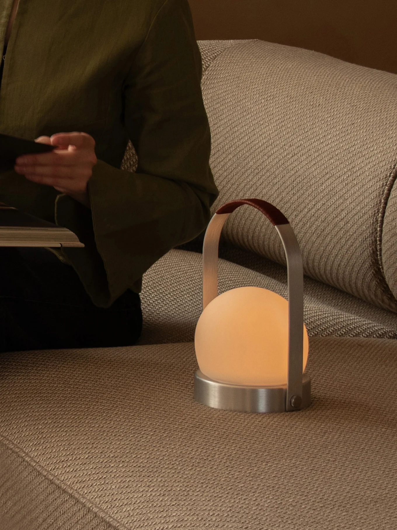 Person liest auf Sofa mit leuchtender Handleuchte mit mattierter Kugel, Metallfuß und Ledergriff.