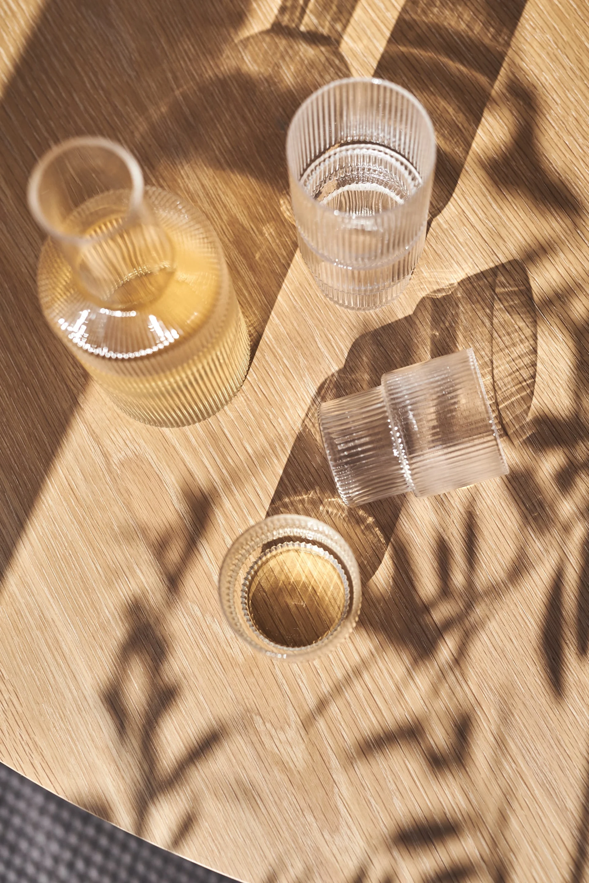 Eine gerippte Glasflasche und zwei gerippte Gläser, eines mit Flüssigkeit, auf einem sonnenbeschienenen Holztisch mit Blattschatten.