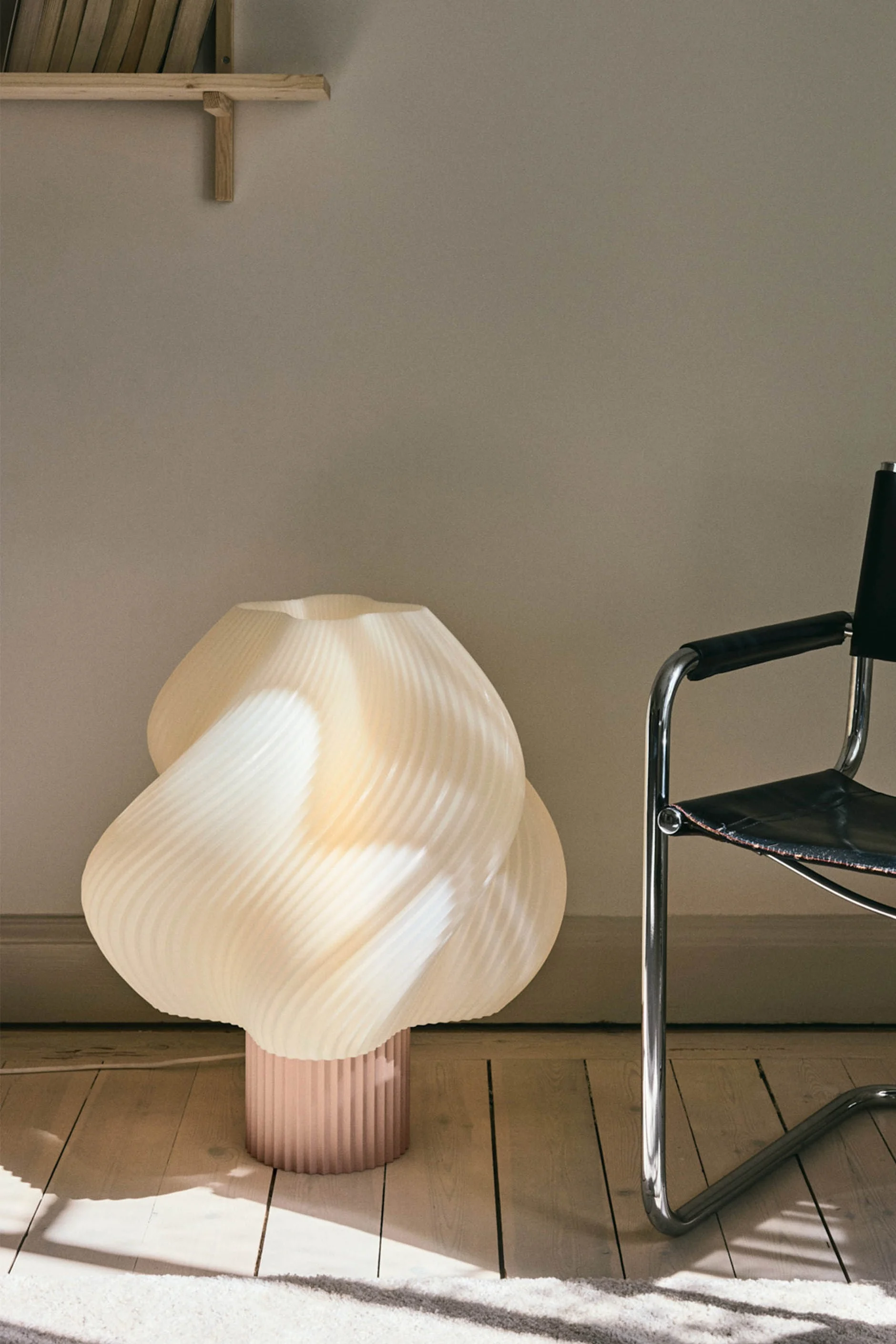 Eine skulpturale, plissierte cremefarbene Stehlampe mit rosafarbenem Sockel steht auf einem Holzfußboden neben einem Stuhl aus schwarzem Leder und Chrom.