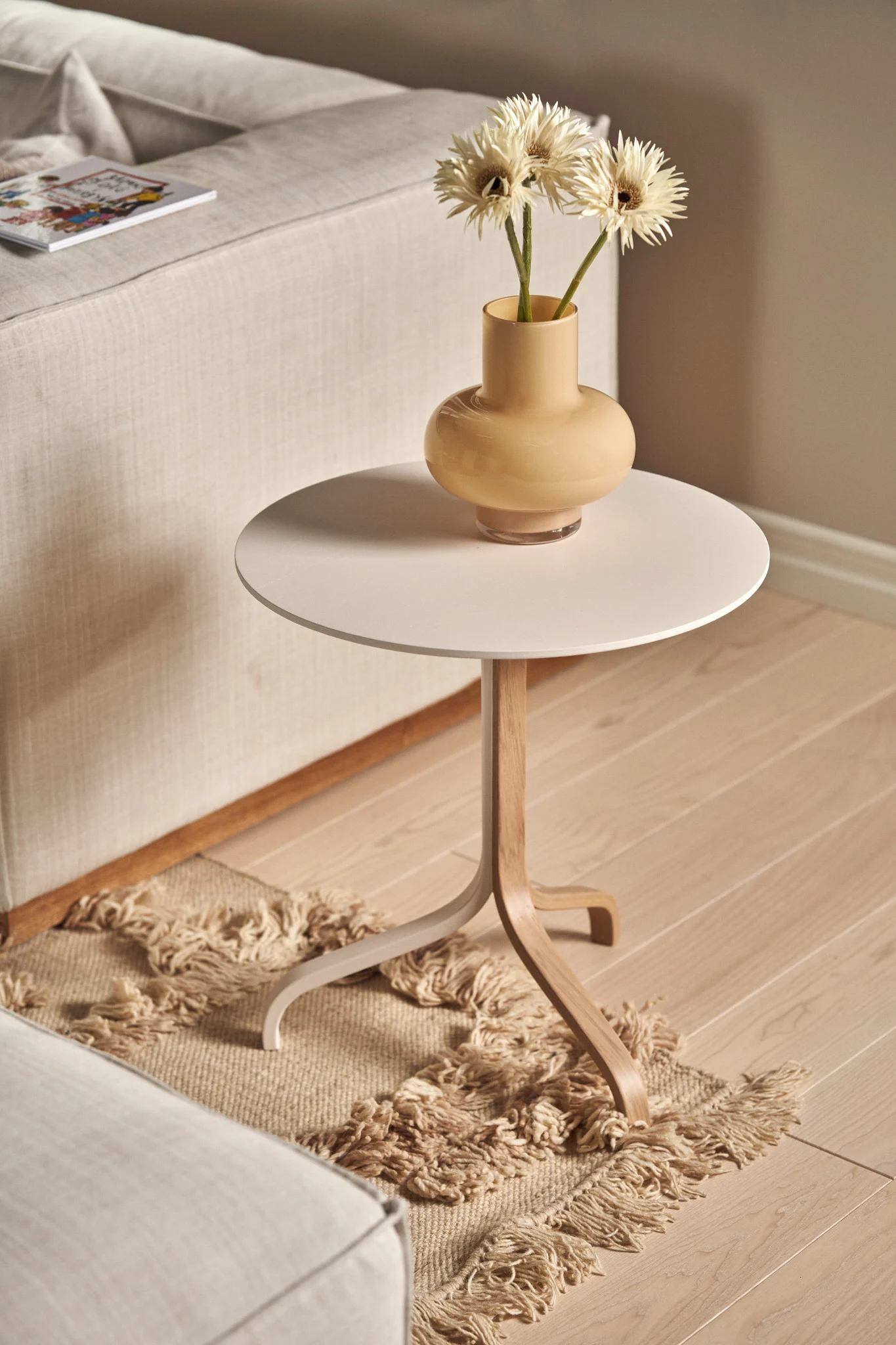 Heller runder Beistelltisch mit Holzfuß, darauf eine Vase mit weißen Blumen, neben einem beigen Sofa und Teppich.