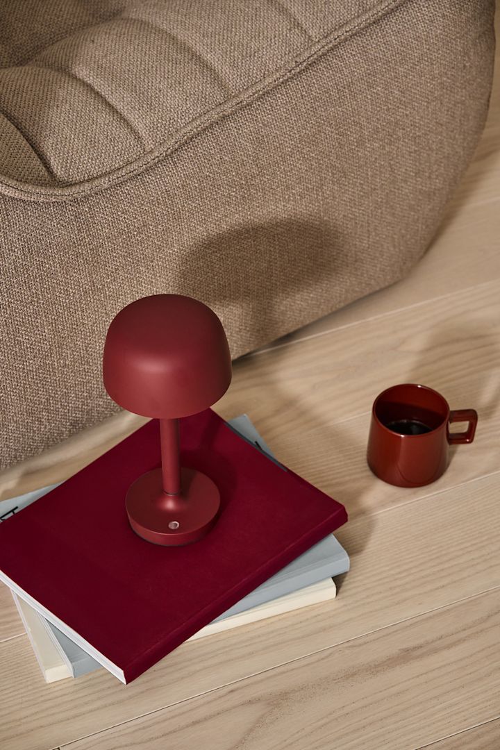 Ein dunkles Rot gehört zu den Wohntrends 2025. Hier steht die burgunderfarbene kabellose Halo-Tischleuchte auf einem Stapel Bücher, daneben die rote Taper-Tasse, beides von Scandi Living.