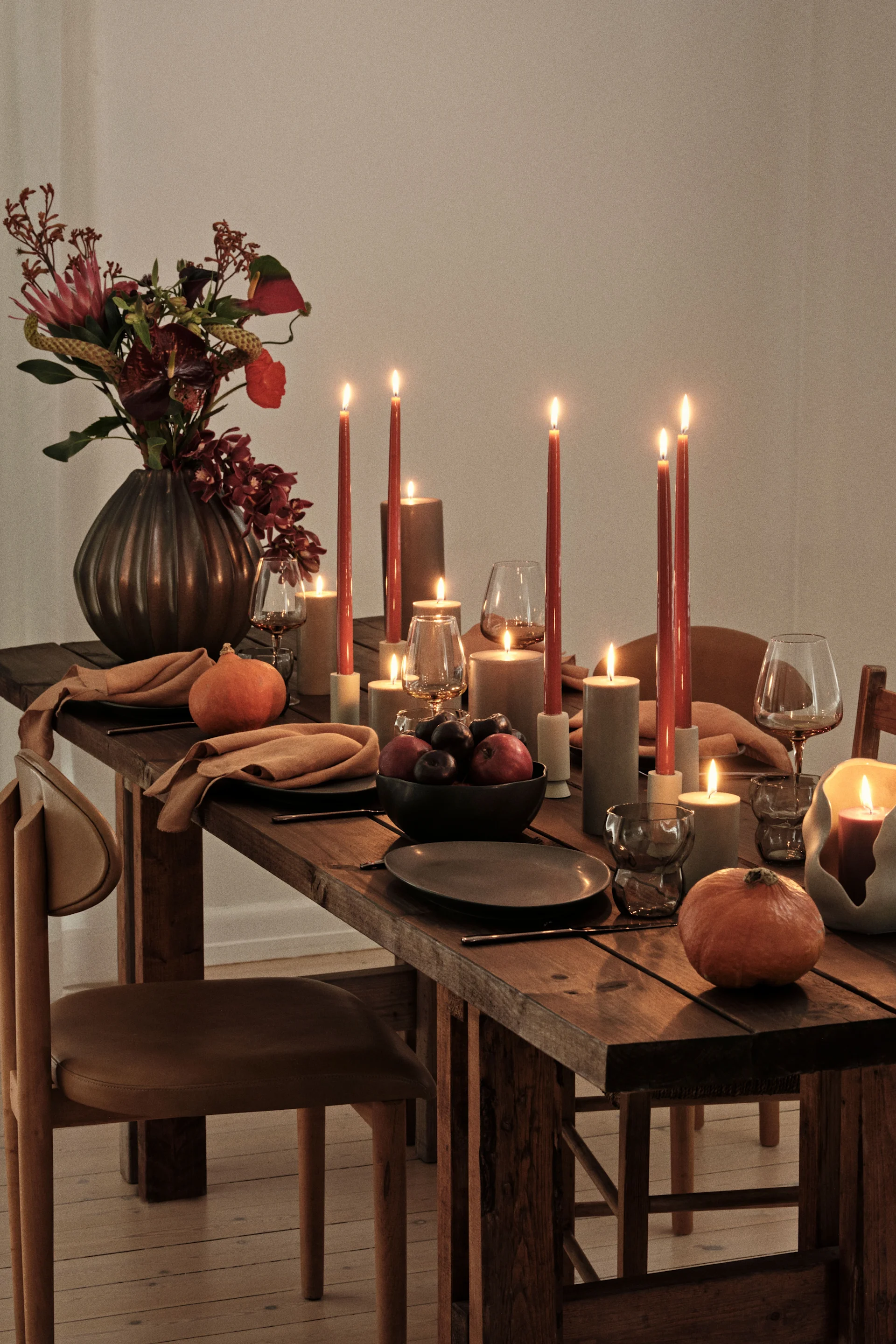 Ein rustikaler Holztisch, gedeckt mit brennenden Kerzen, dunklen Tellern, beigefarbenen Servietten, einem Blumenarrangement in einer dunklen Vase und Kürbissen, schafft ein warmes Ambiente.