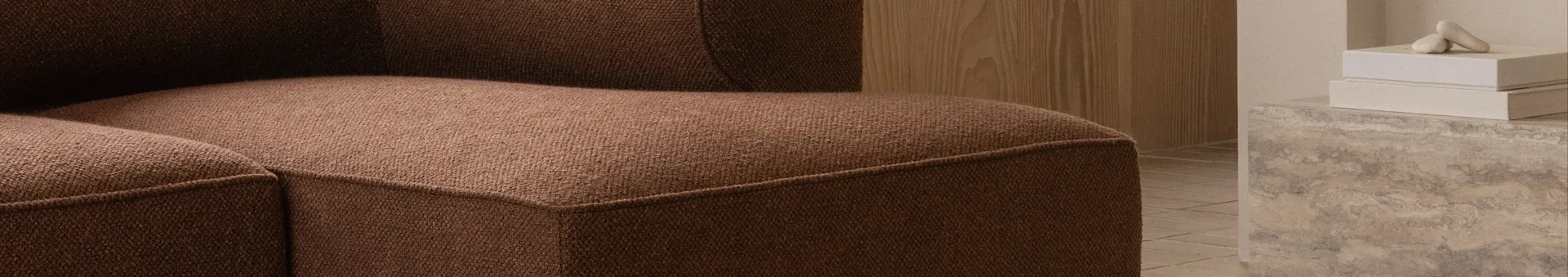 Nahaufnahme eines braunen strukturierten Sofas, mit einer Holzwand und Deko-Objekten auf einer Betonfläche im Hintergrund.