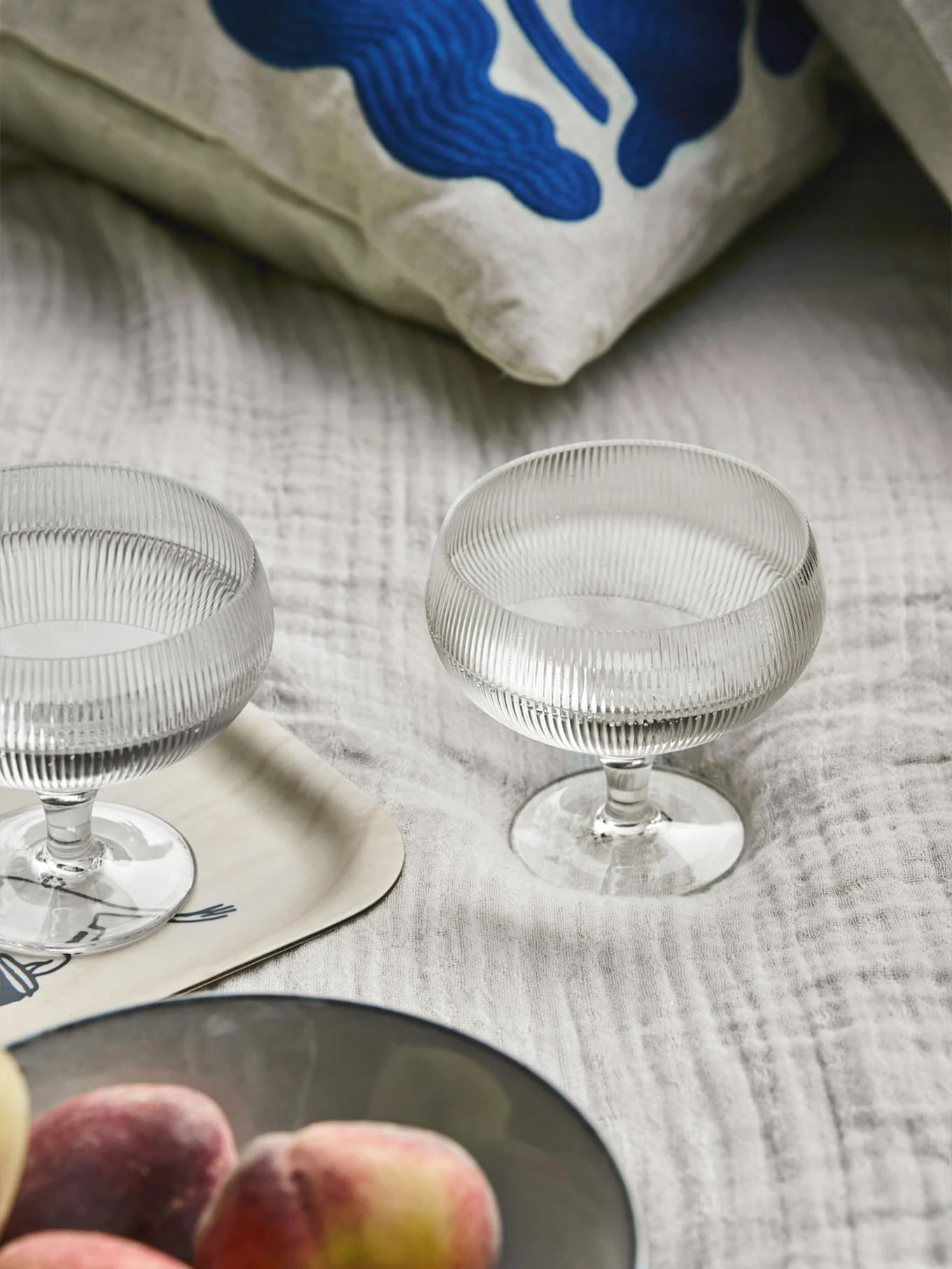 Zwei gerippte Glasdessertschalen, ein Kissen mit blauer abstrakter Kunst, ein Holztablett und Pfirsiche auf hellem Stoff.