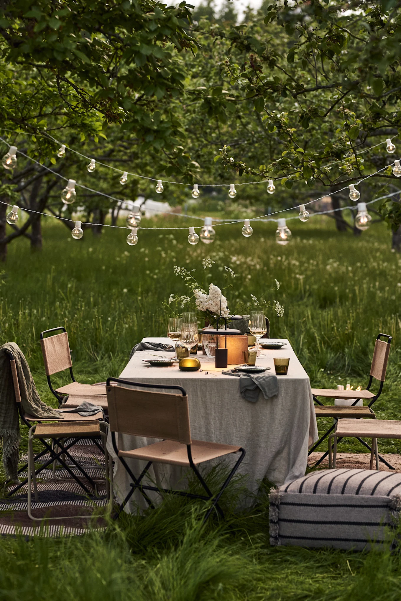 Esstisch im Freien mit weißen Blumen, Laterne, Korbstühlen und Lichterketten im grünen Obstgarten.