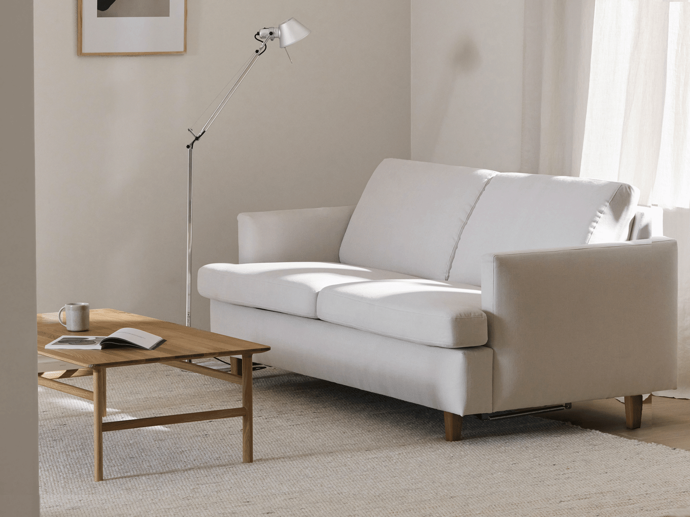 Ein minimalistisches Wohnzimmer mit einem hellgrauen Zweisitzer-Sofa, einem Holztisch, einer modernen Stehlampe und transparenten Vorhängen.