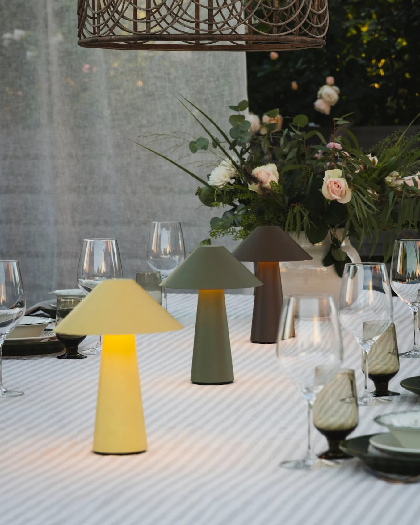 Esstisch im Freien mit drei beleuchteten Pilzlampen, Blumenarrangement und einer Korblampe.