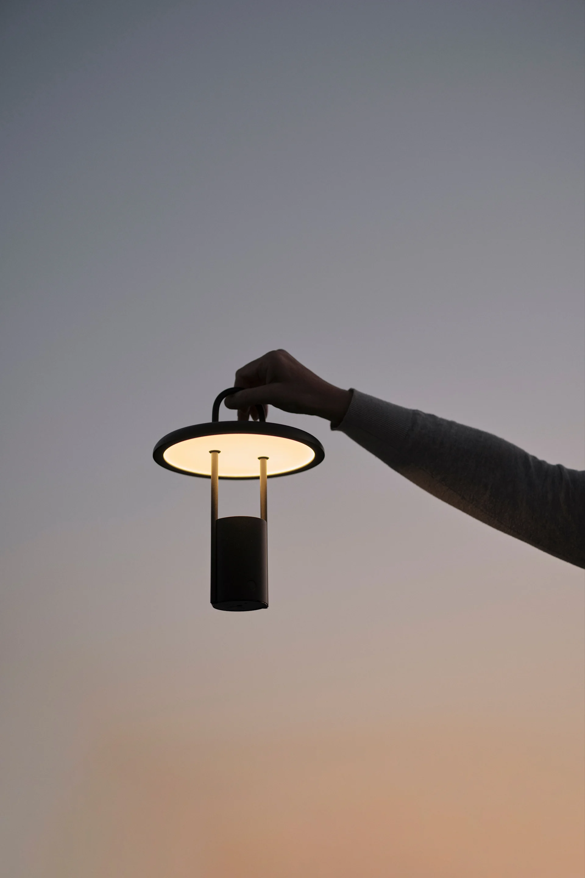 Eine Hand hält eine leuchtende Akku-Tischleuchte vor einem farbverlaufenden Dämmerungshimmel.