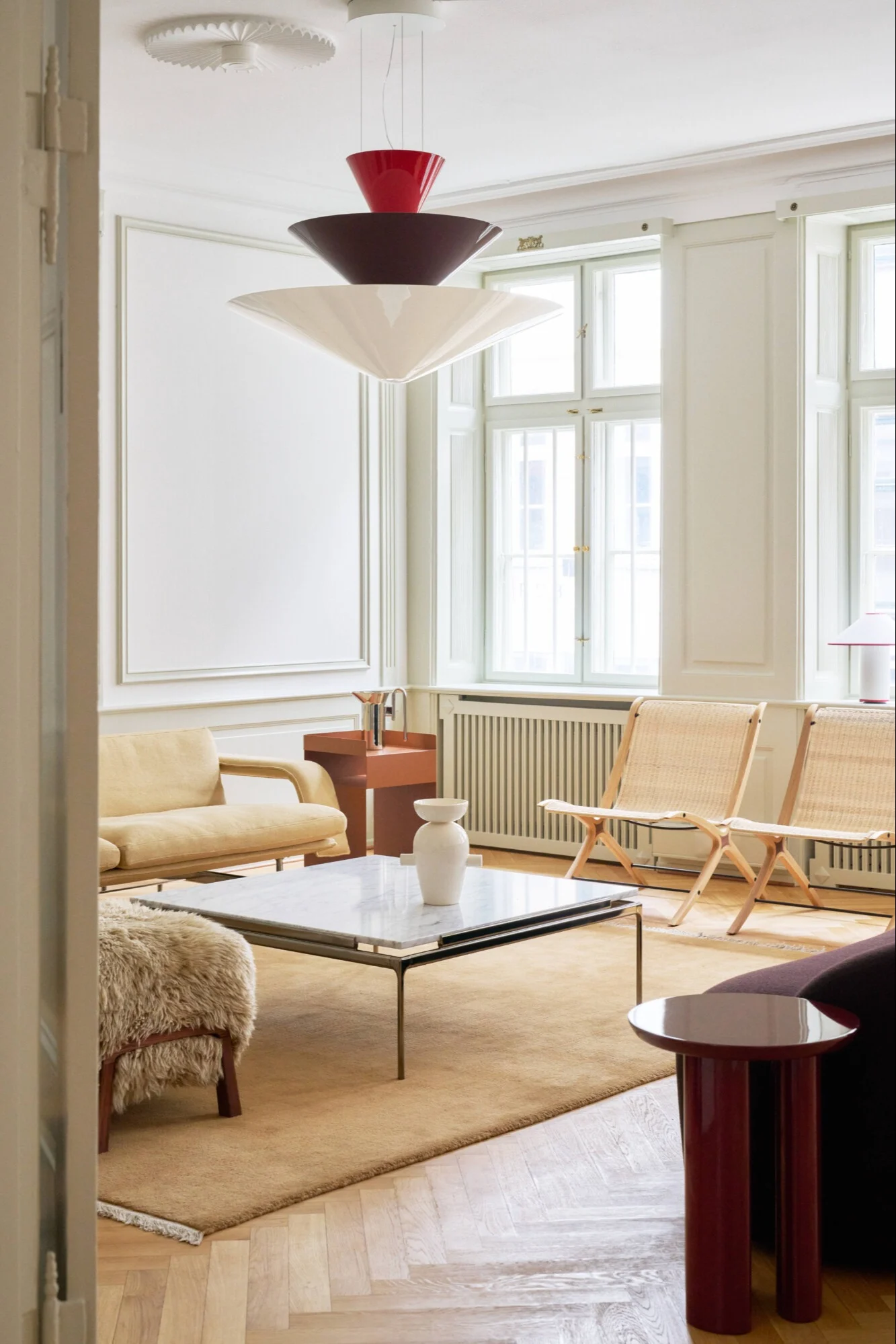 Ein modernes Wohnzimmer mit einer gestuften Pendelleuchte, hellgelbem Sofa, Korbstühlen und Marmor-Couchtisch.