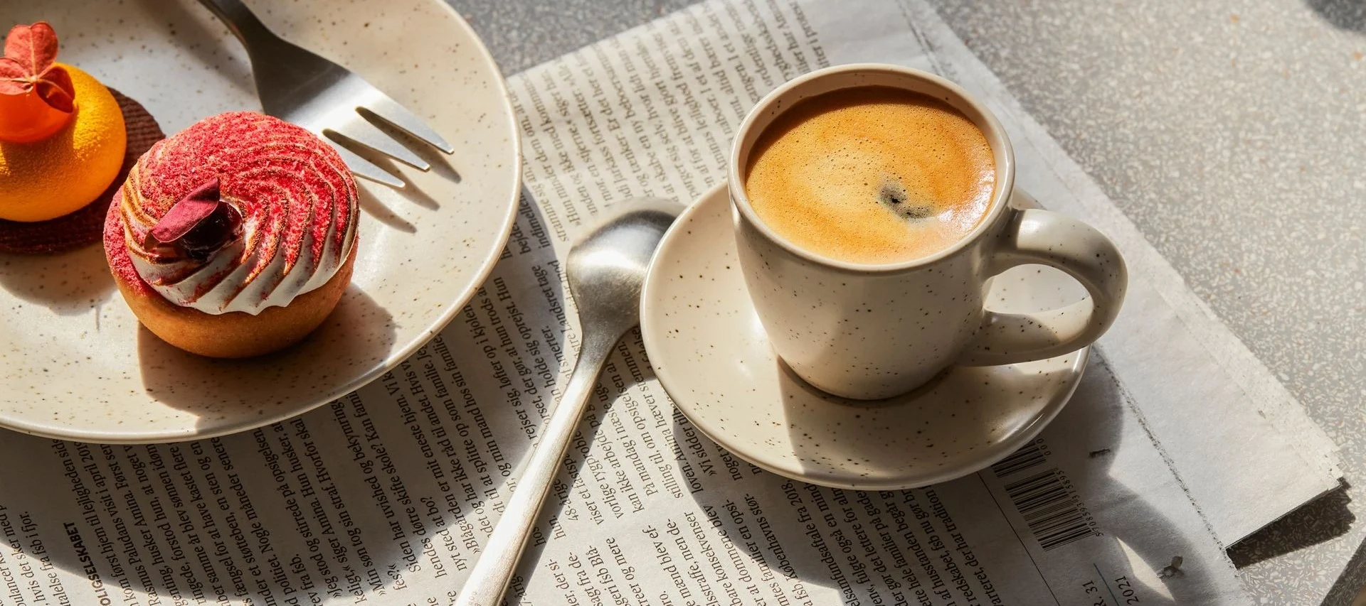 Kaffeetasse und zwei Gebäckstücke, eine rote Tarte und eine orangefarbene, auf einer Zeitung.