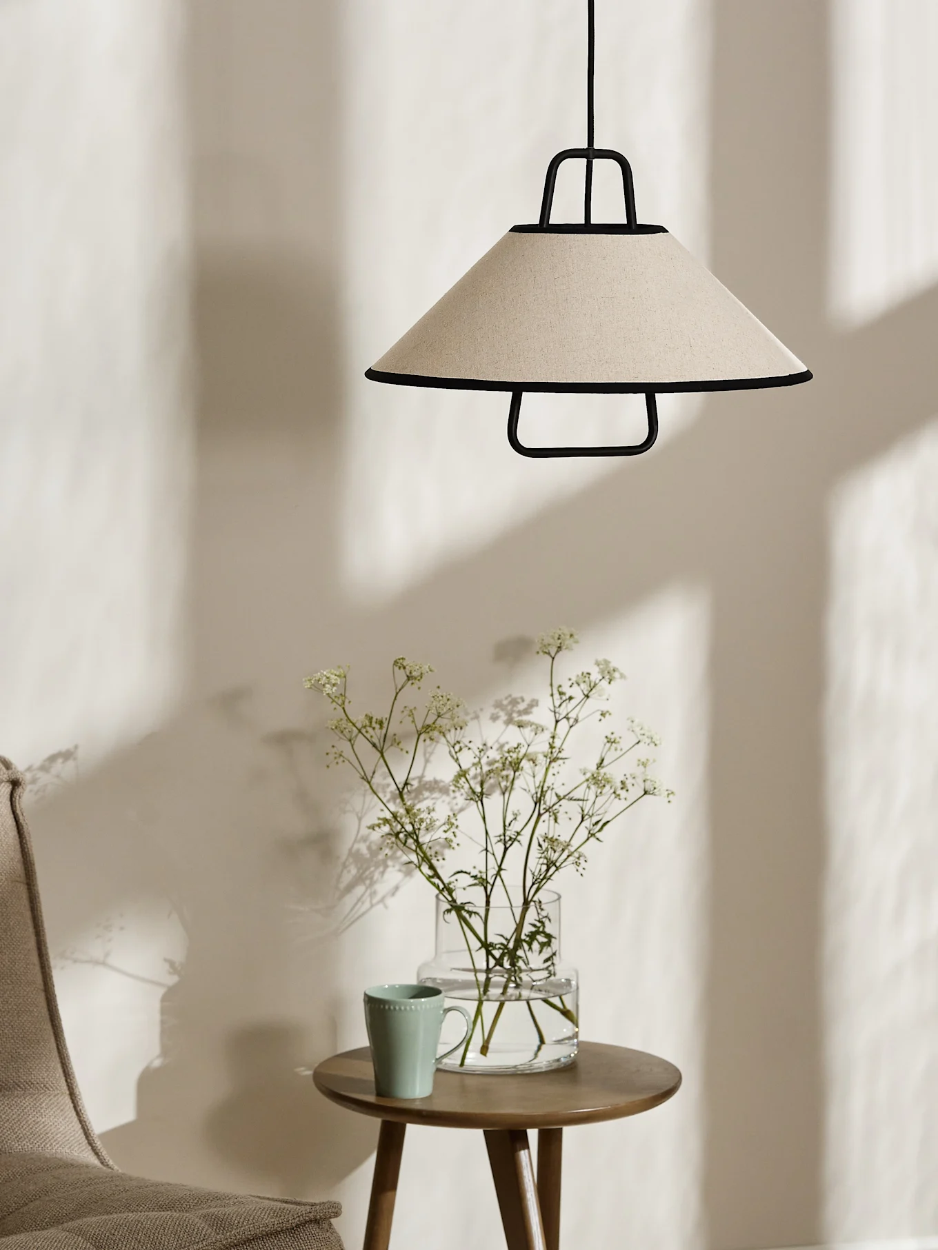 Eine konische cremefarbene Pendelleuchte mit schwarzem Rand hängt über einem Holznebentisch mit einer Vase weißer Blumen und einer hellgrünen Tasse.