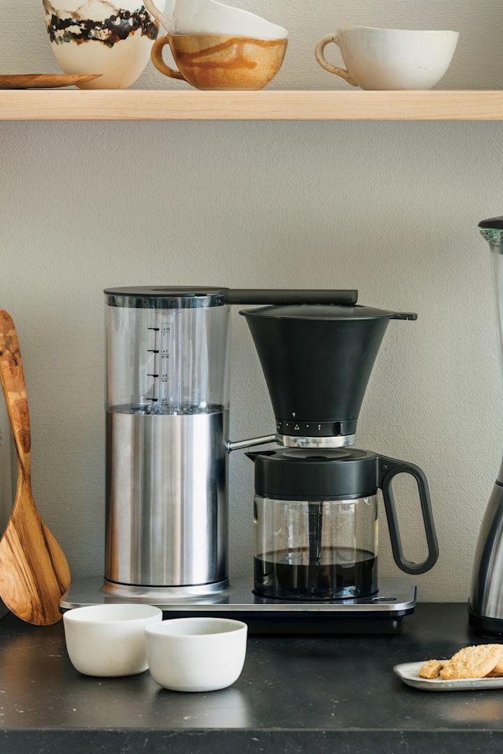 Für Ihre Kaffeebar zu Hause benötigen Sie auch die richtigen Geräte. Hier sehen Sie eine klassische Filterkaffeemaschine von Wilfa auf einer Küchenanrichte stehen, darüber ein Regal mit Schüsseln und einem Toaster.