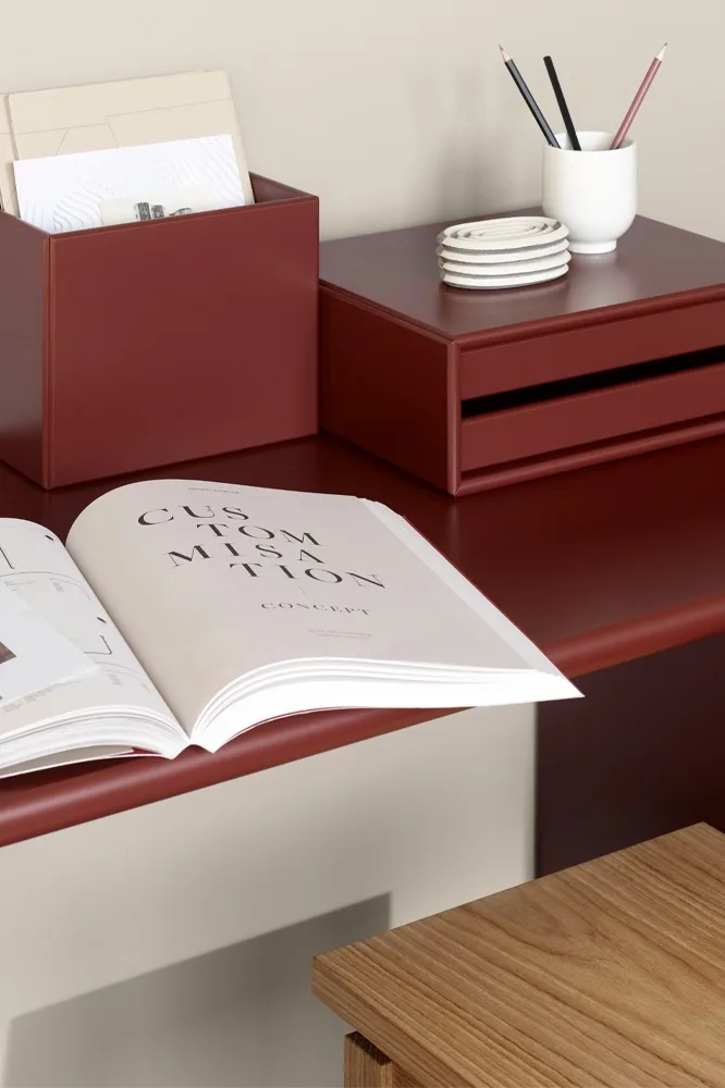 Ein rotbrauner Schreibtisch mit einer Aktenbox, einem Schubladenschrank, einem Stifthalter, Untersetzern und einem offenen Buch. Ein Holzhocker ist teilweise sichtbar.