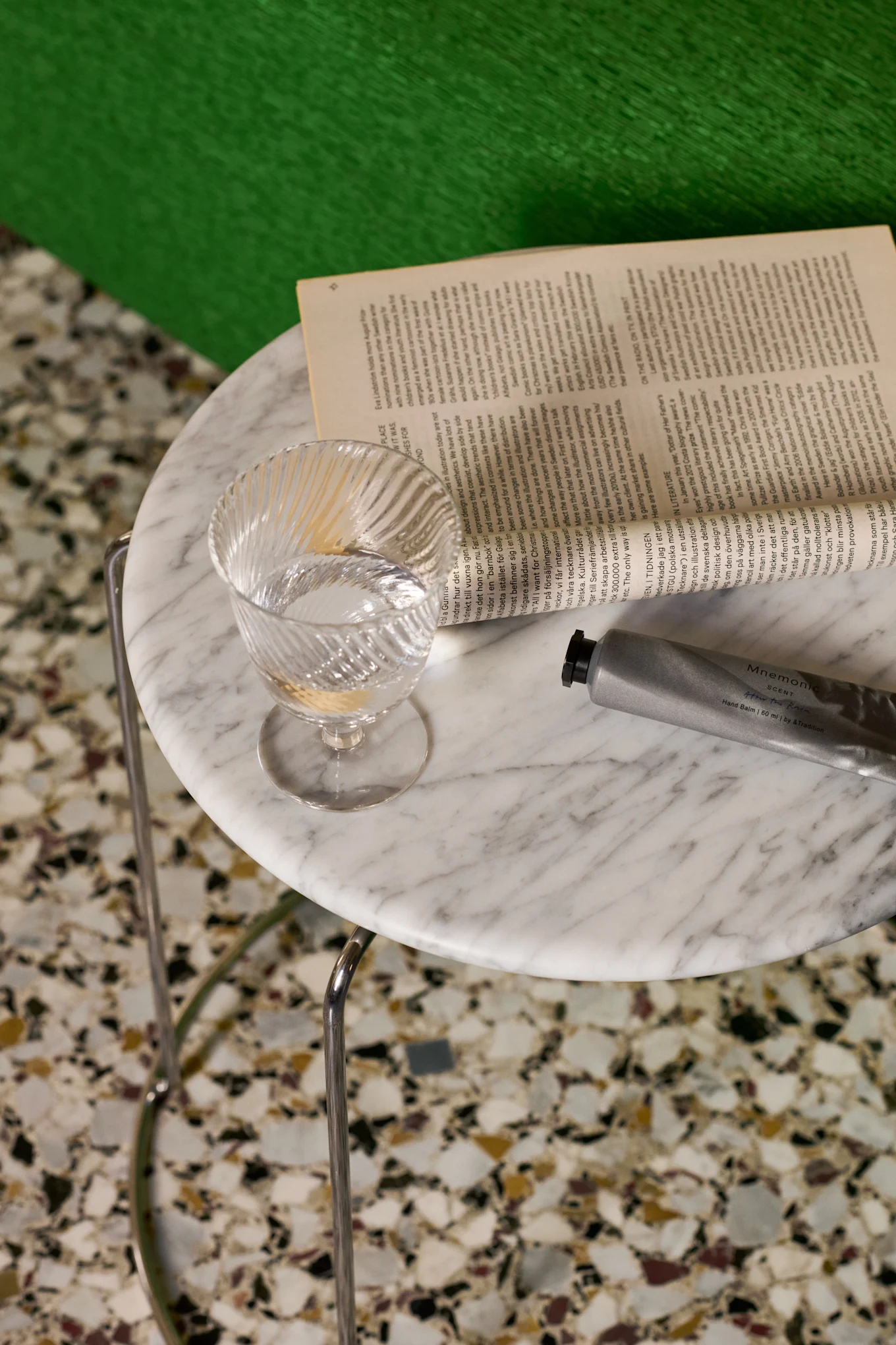 Ein klares, geriffeltes Wasserglas, ein offenes Buch und eine silberne Handbalsamtube auf einem Marmortisch.