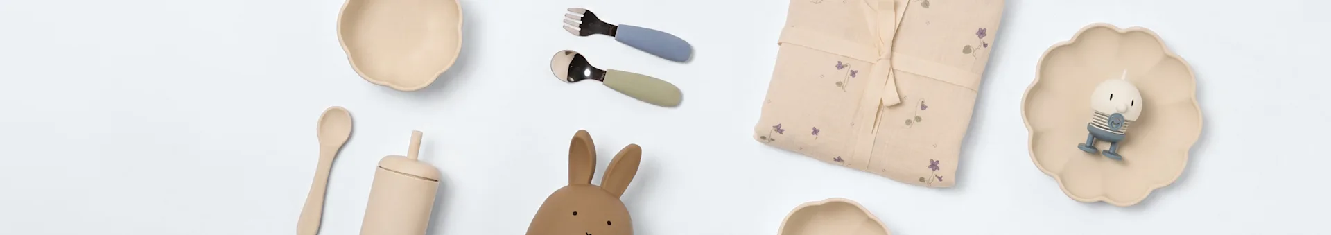 Babyartikel auf Weiß: zwei cremefarbene Schalen, blaues und grünes Besteck, florales Tuch, Hasenspielzeug, Holzlöffel, beiger Becher und kleine Figur auf wellenförmigem Teller.