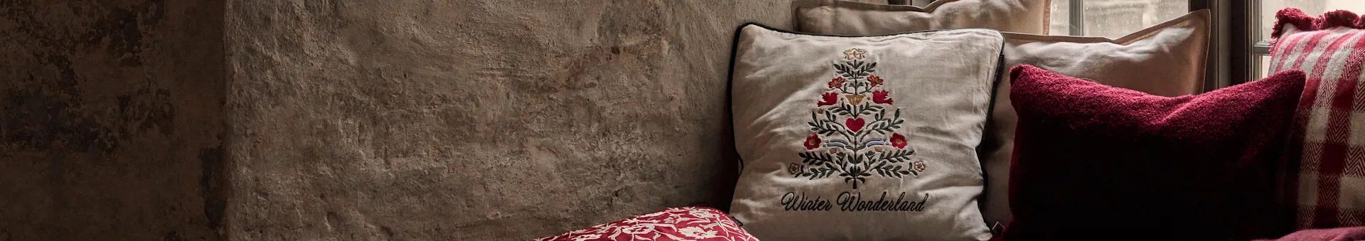 Kissen, darunter ein besticktes Weihnachtsbaumdesign mit "Winter Wonderland"-Text, ein dunkelrotes Kissen und ein rot-weiß kariertes Kissen.