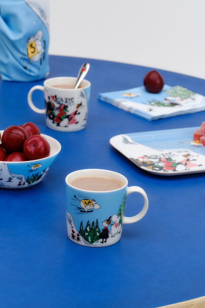 Die Mumin-Tasse Winter 2024, "Skispringen", steht auf einem in Blau gedeckten Tisch.