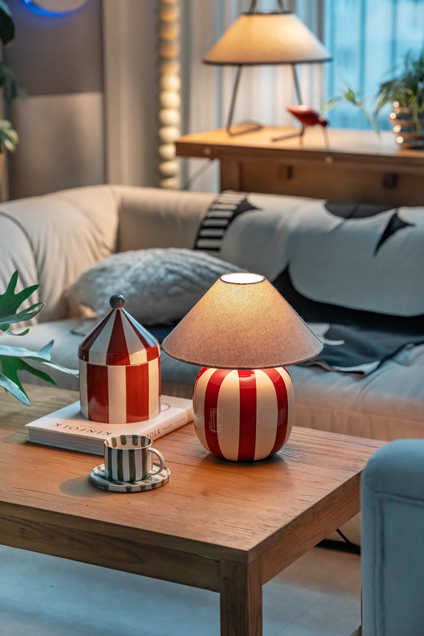 Ein gemütliches Wohnzimmer mit einer rot-weiß gestreiften Lampe, passendem Behälter und einer grün gestreiften Tasse auf einem Couchtisch aus Holz.