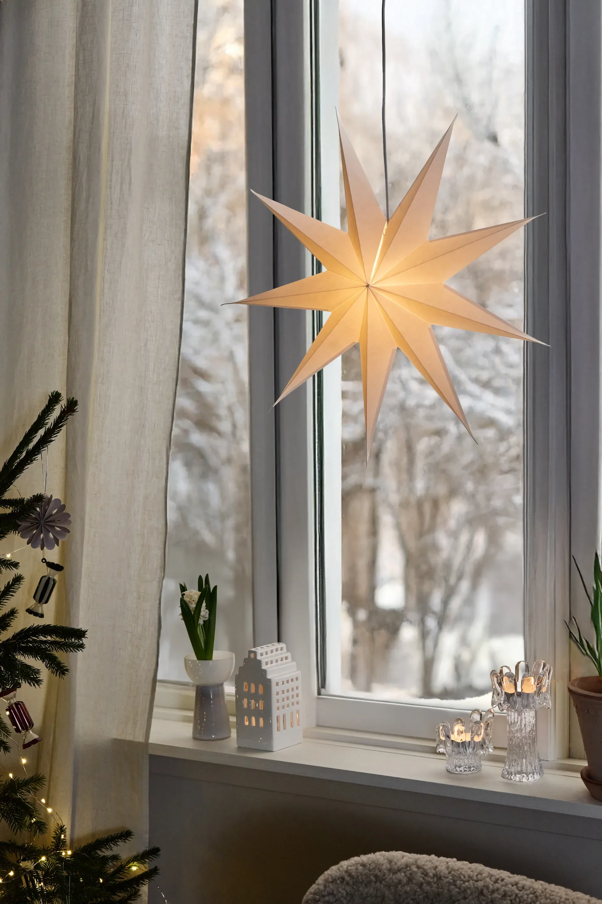 Fenster dekoriert für den Winter mit leuchtendem Papierstern, Keramikhaus, brennenden Kerzen und Weihnachtsbaum.
