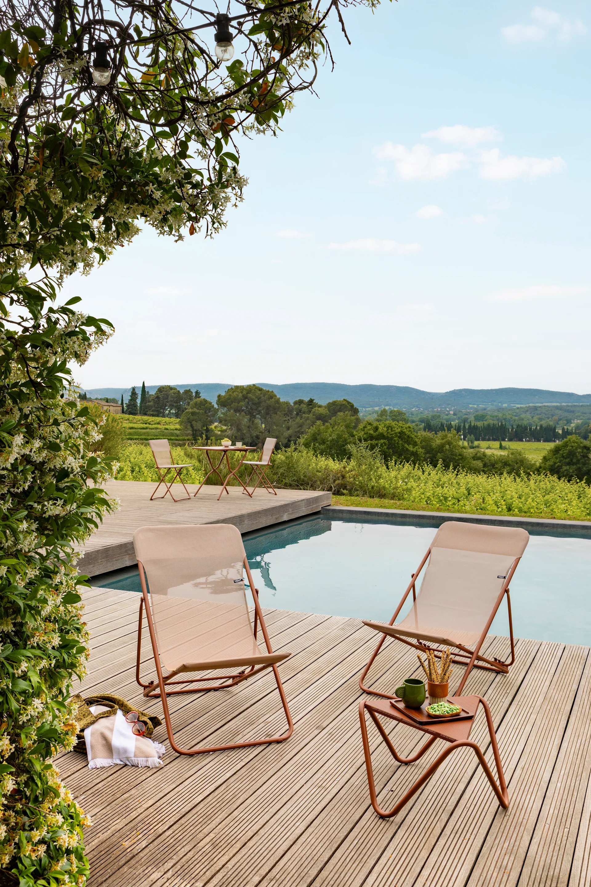 Zwei hellbraune Outdoor-Liegestühle und ein Beistelltisch auf einer Holzterrasse am Pool, mit blühenden Ranken und Weinbergblick.
