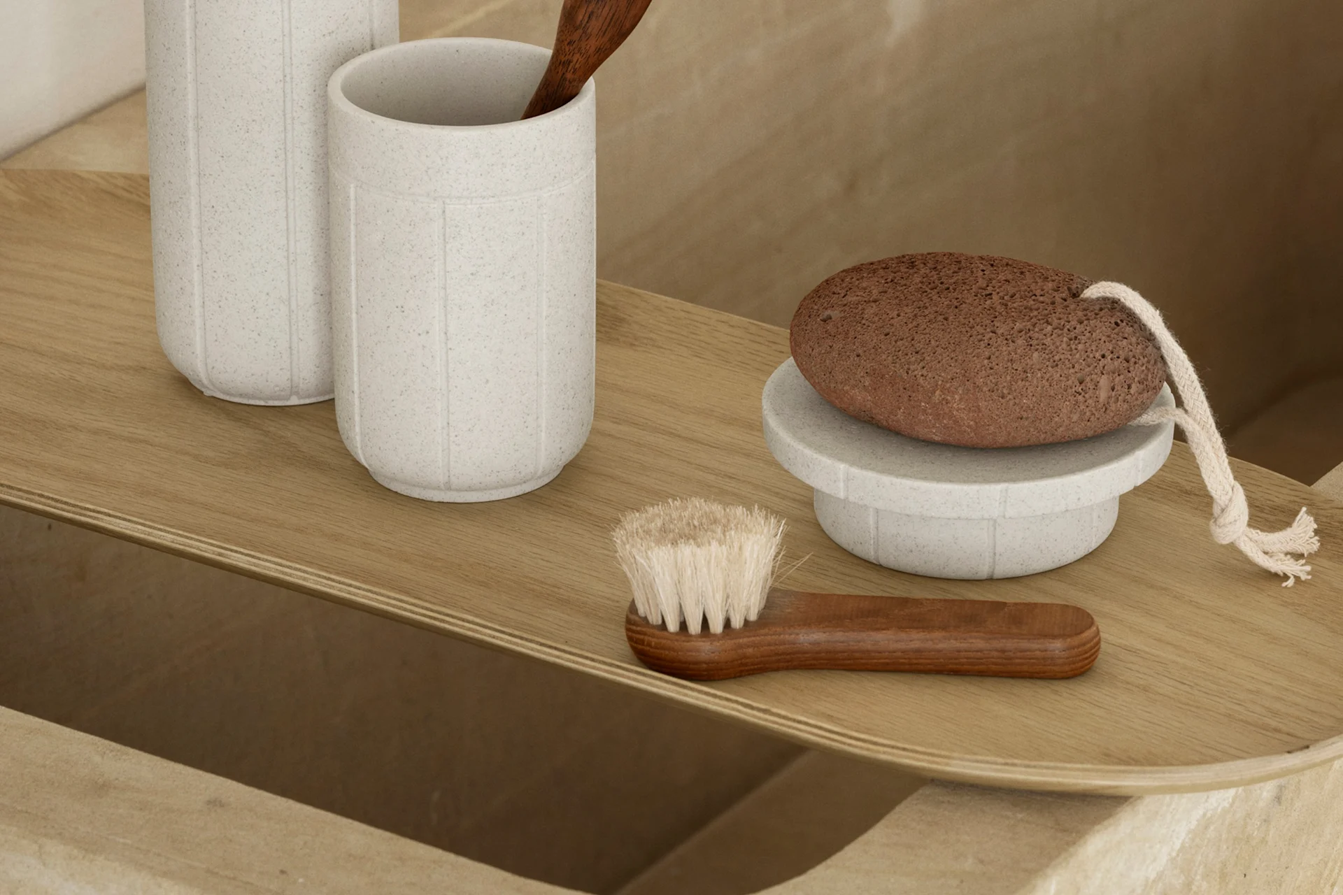 Ein helles Holztablett mit Badaccessoires: zwei gesprenkelte graue Becher, ein Holzlöffel, ein brauner Bimsstein auf einem Ständer und eine Holzbürste.
