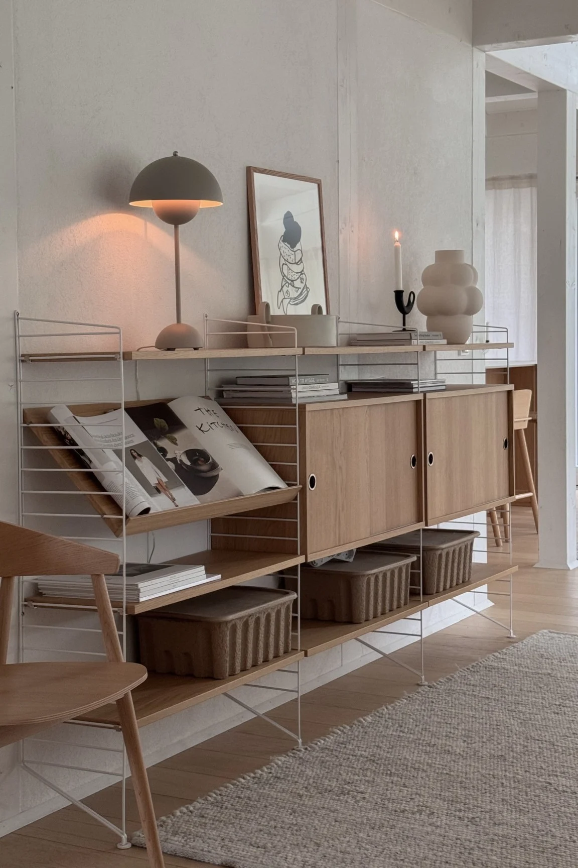 Modernes Regal mit Zeitschriften, Büchern, Körben, Lampe, Kunst, Kerze und Vase in neutralem Raum.