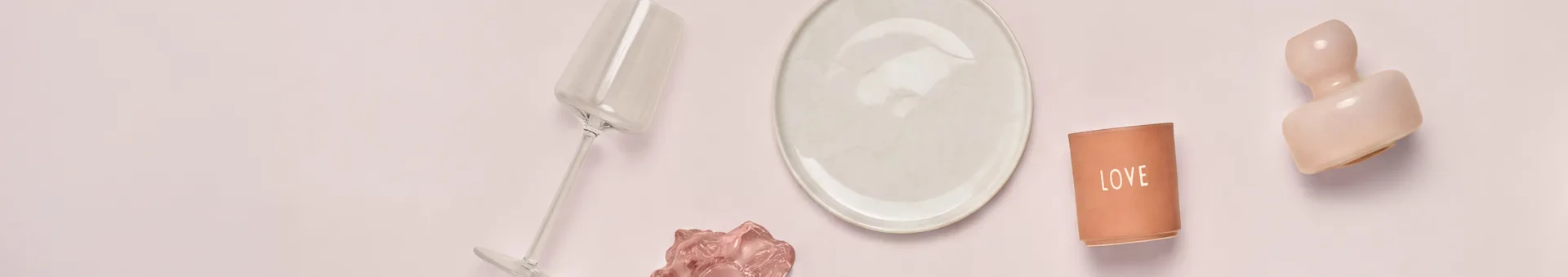 Eine Draufsicht auf verschiedene Dekorations- und Essgegenstände auf einer hellrosa Oberfläche. Von links nach rechts: ein gekipptes klares Glas, ein cremefarbener Teller, ein terrakottafarbener "LOVE" Kerzenhalter und eine rosa abstrakte Skulptur. Ein kleineres rosa Wellenobjekt ist unter dem Glas.
