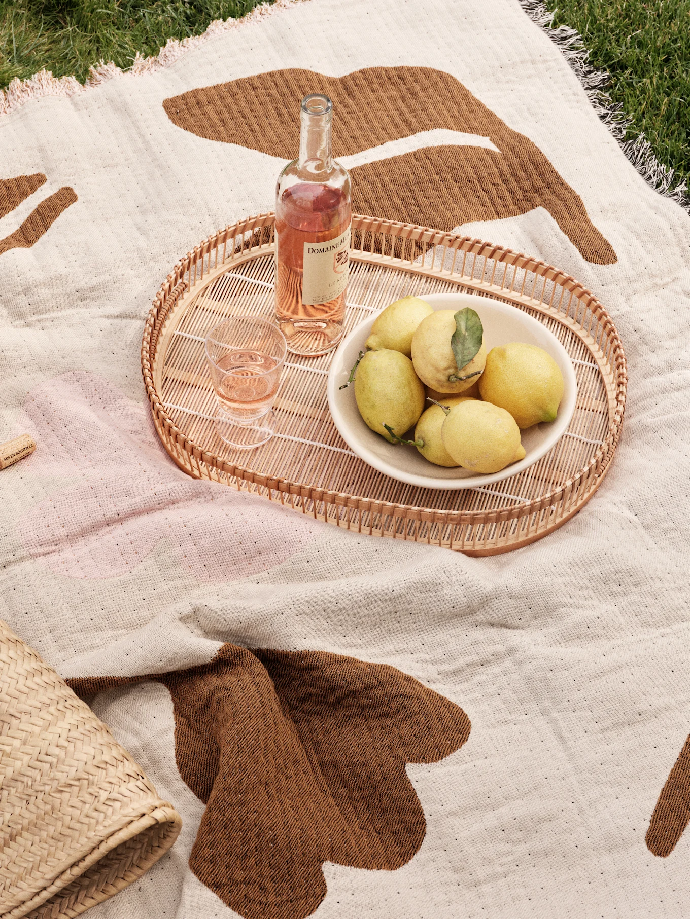 Picknickdecke auf Gras mit Rattan-Tablett, Roséwein, Glas und einer Schale Zitronen.